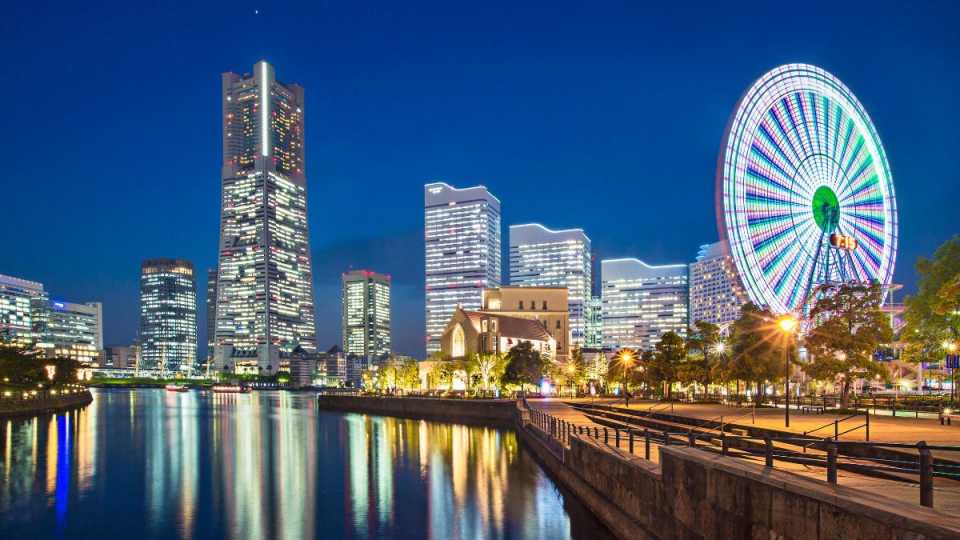 Beleuchtete Skyline einer Stadt bei Nacht mit Hochhäusern und einem großen Riesenrad, das sich im Wasser spiegelt